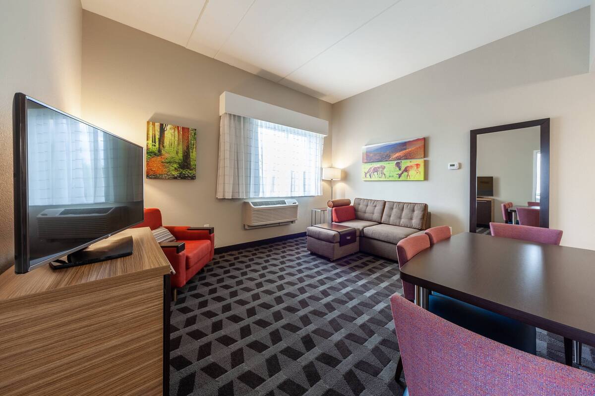 A hotel room with a flat-screen TV, a sofa, a dining table, and a carpeted floor.