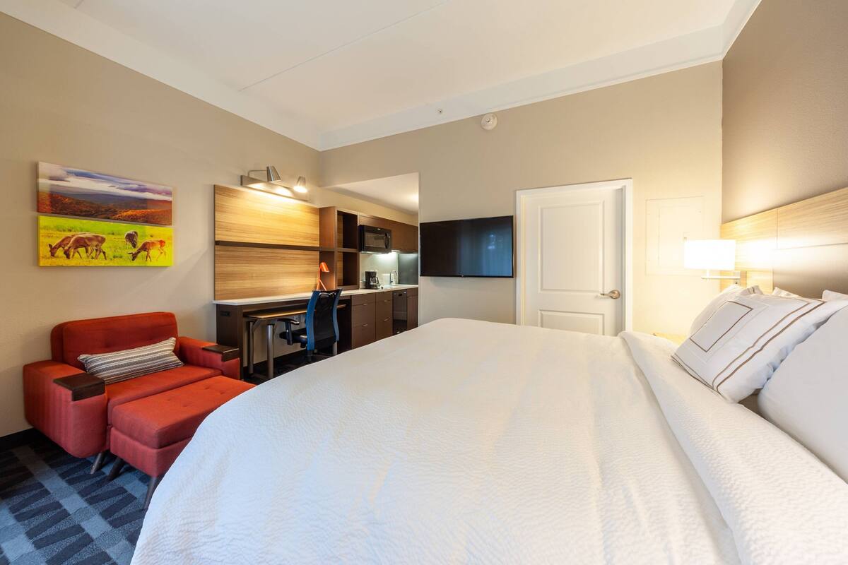 A hotel room with a large bed, a red armchair, a desk with a computer, a flat-screen TV, and a painting on the wall.
