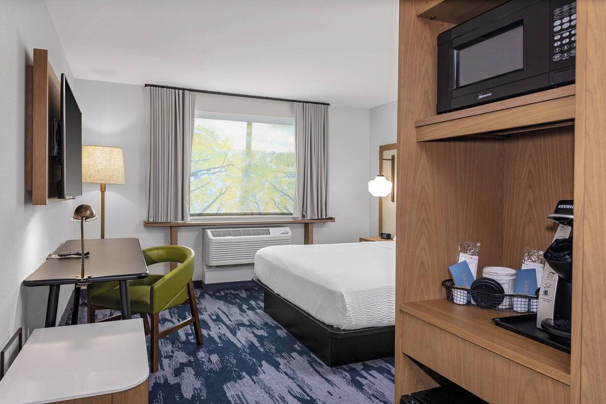 Ein Hotelzimmer mit einem Bett, einem Schreibtisch mit Stuhl, einem Fernseher, einer Lampe, einem Fenster mit Vorhängen, einer Klimaanlage, einer Mikrowelle und einer Kaffeemaschine.