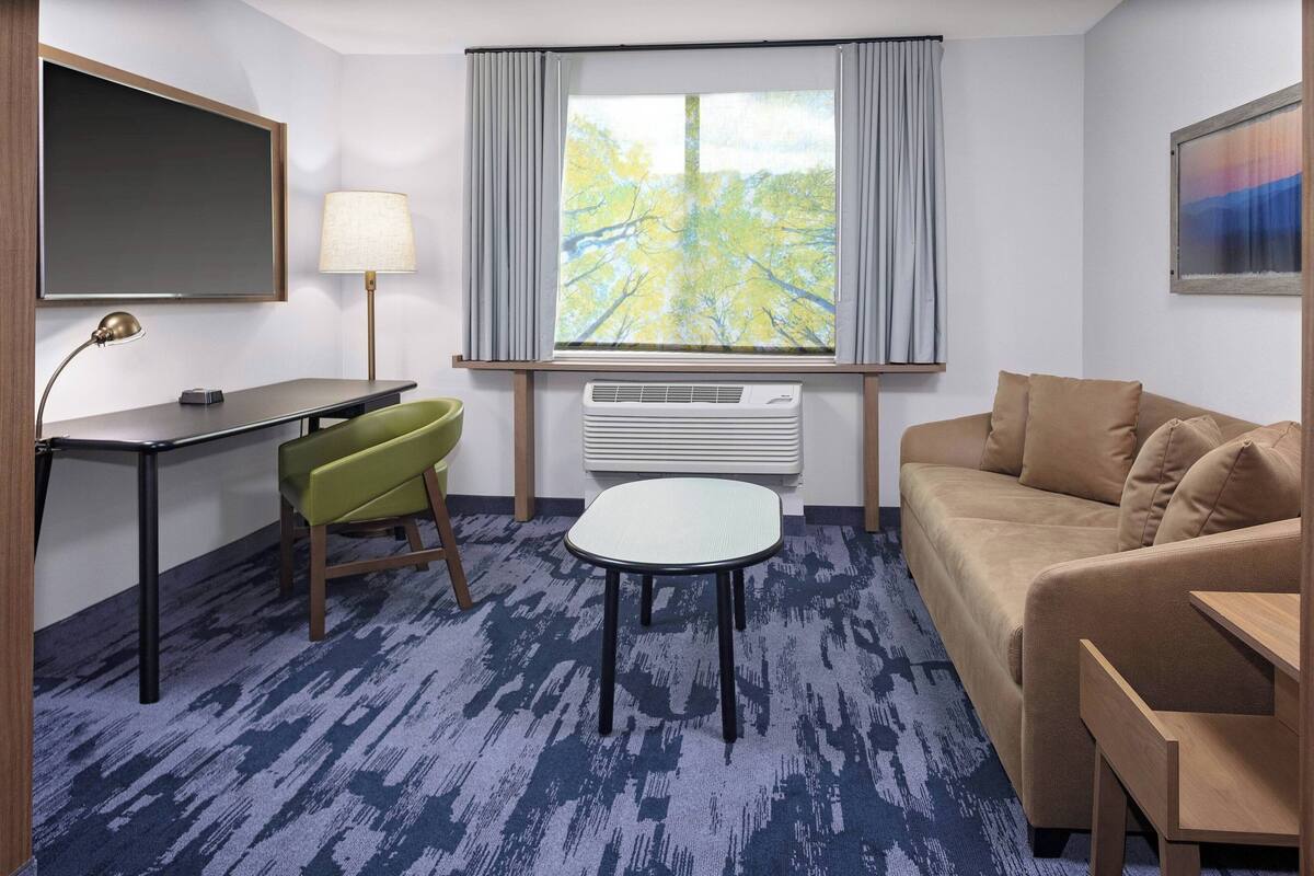 Ein Hotelzimmer mit Schreibtisch, einem grünen Sessel, einem beigen Sofa, einem kleinen Tisch, einem großen Fenster mit Vorhängen, einem Flachbildfernseher, einer Lampe und einer Klimaanlage.