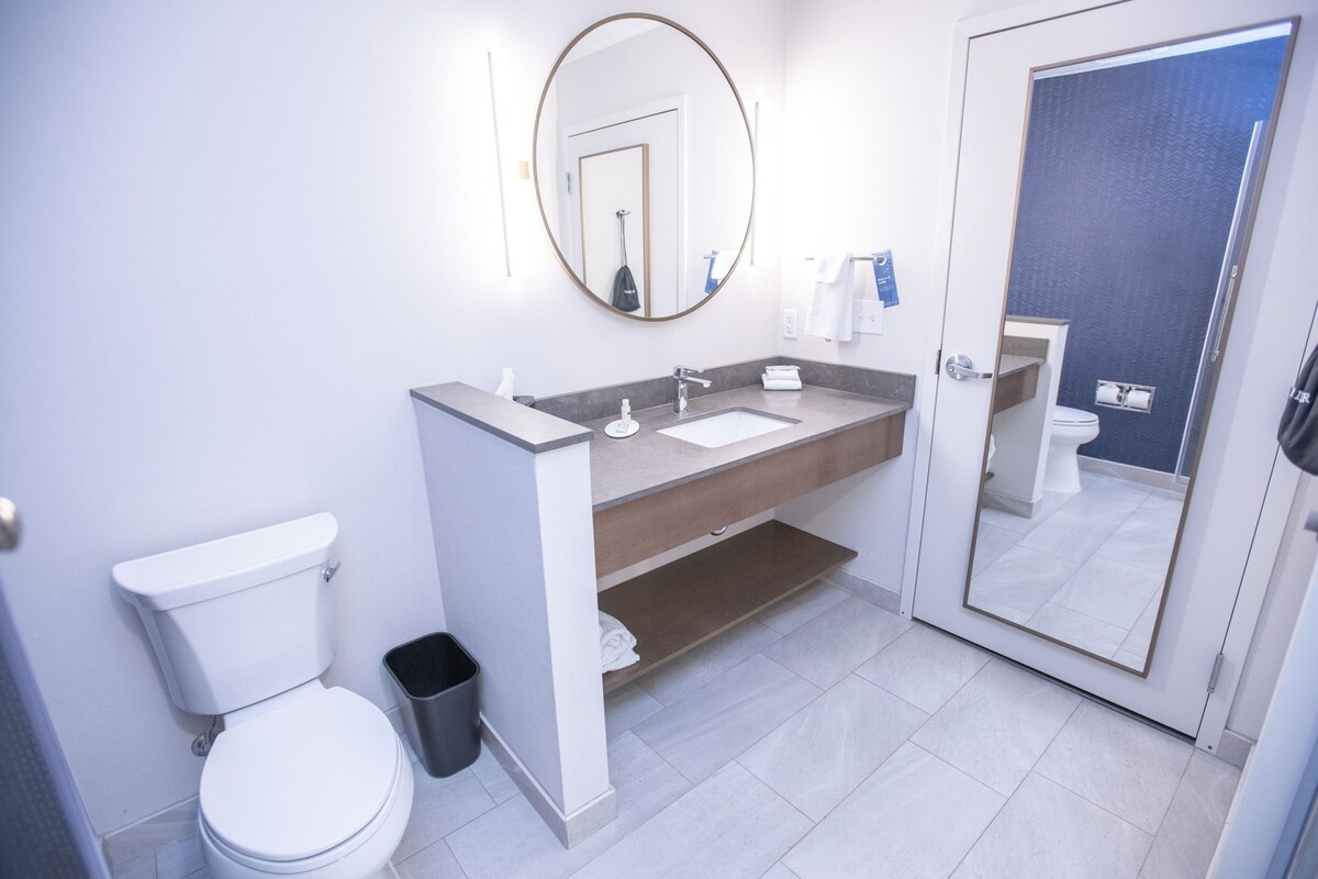 Un baño moderno con inodoro blanco, lavamanos con espejo y zona de ducha visible a través de un vidrio.