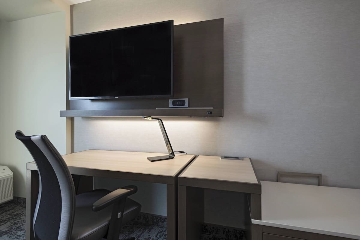 A modern hotel room with a flat-screen TV mounted on the wall, a desk with a lamp, and a chair.
