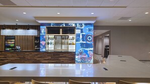 A modern bar area with a digital menu display, a seating area with a long table and chairs, and a wall with a colorful abstract design.