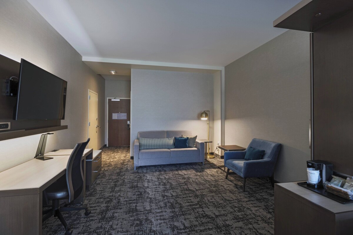 Habitación de hotel con escritorio, sofá, sillón, televisor, máquina de café y lámpara.