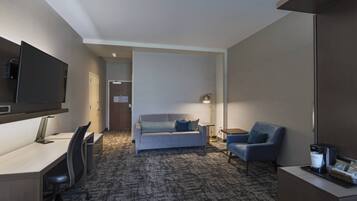 A hotel room with a desk, a sofa, a chair, a TV, a coffee machine, and a lamp.