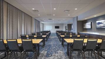 A conference room with rows of tables and chairs, a projector screen, and a long counter with a flat-screen TV.