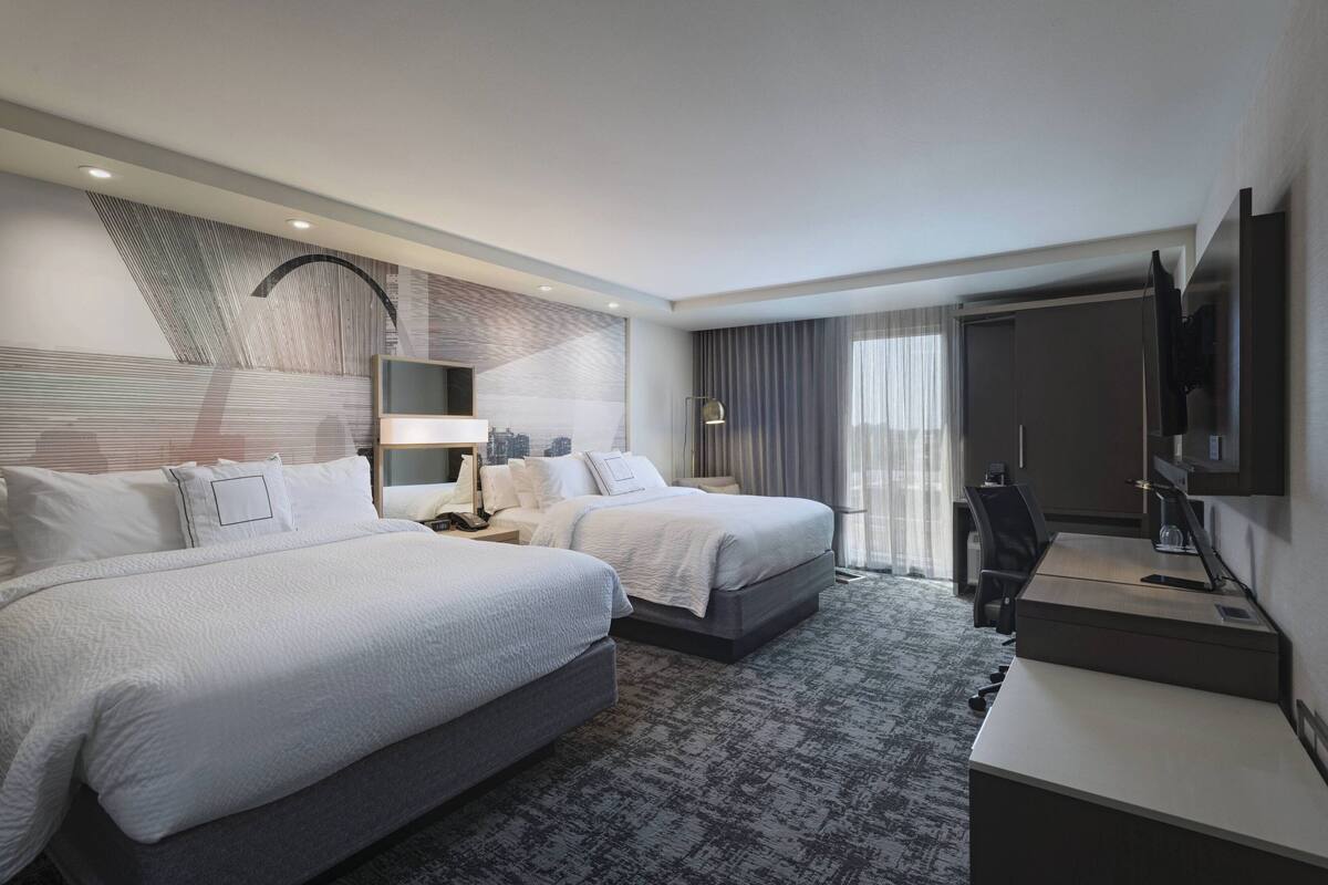 A modern hotel room with two beds, a desk, a TV, and a large window with curtains.