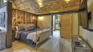 Une chambre avec un plafond en bois, un lit recouvert d’une literie à motifs, une table de chevet ornée d’une plante et une porte donnant sur une autre pièce.