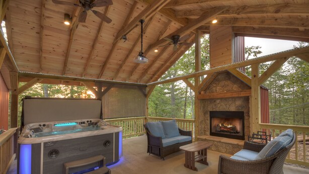 Une terrasse en bois avec un jacuzzi, deux canapés en osier, une petite table et une cheminée en pierre.