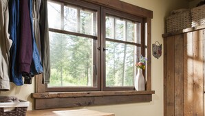 Una habitación rústica con una mesa de madera, cestas, un tapete rojo y un ventanal con vistas a árboles.