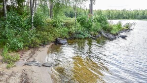 A sandy shoreline with rocks and dense vegetation beside a calm body of water. - Sieralahden lomakylä, mökki 2 by Interhome (Savonlinna)