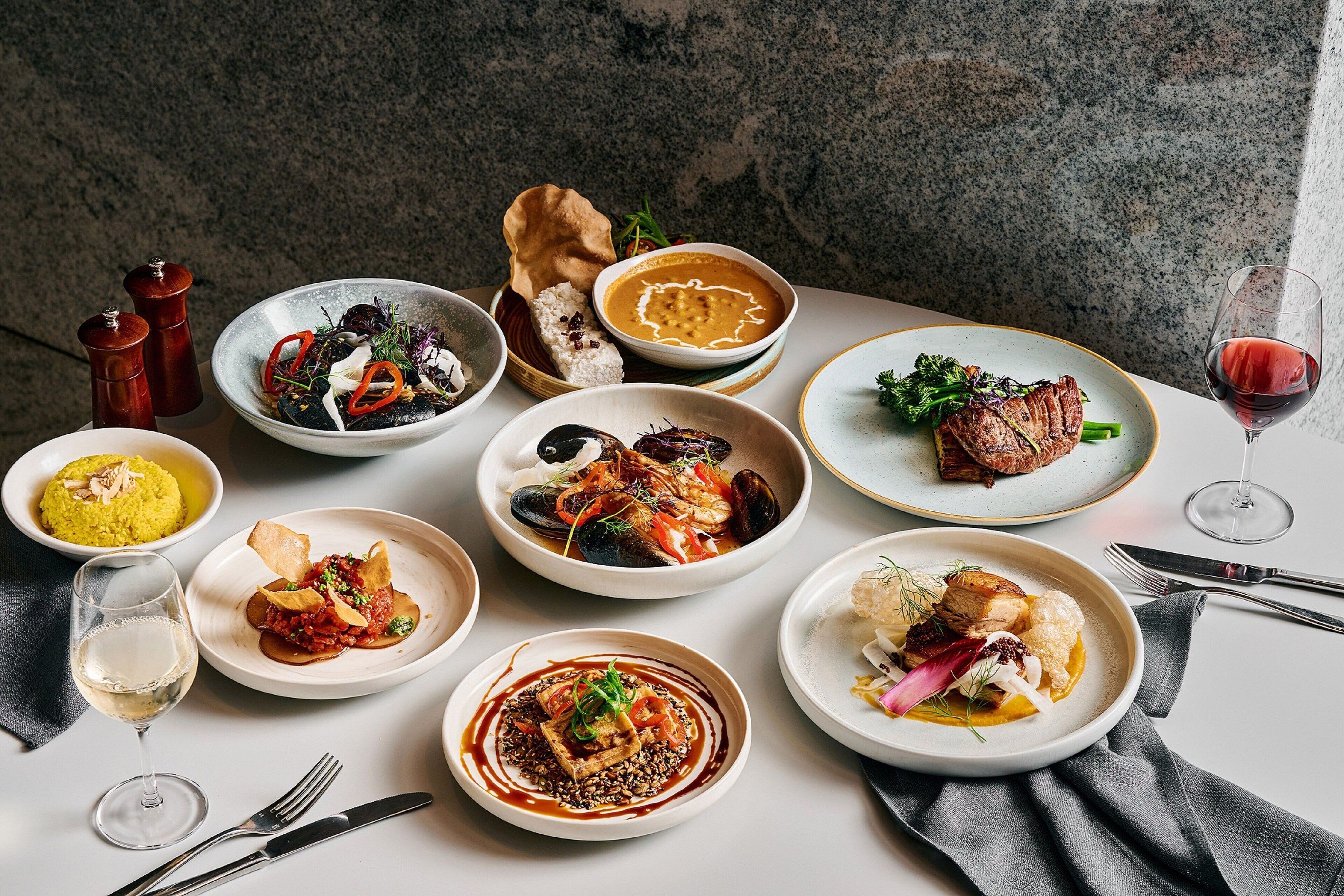 A variety of dishes on a table, including seafood, meat, and vegetables, with a wine glass and cutlery.