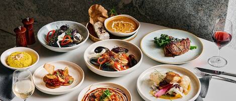 A variety of dishes on a table, including seafood, meat, and vegetables, with a wine glass and cutlery.