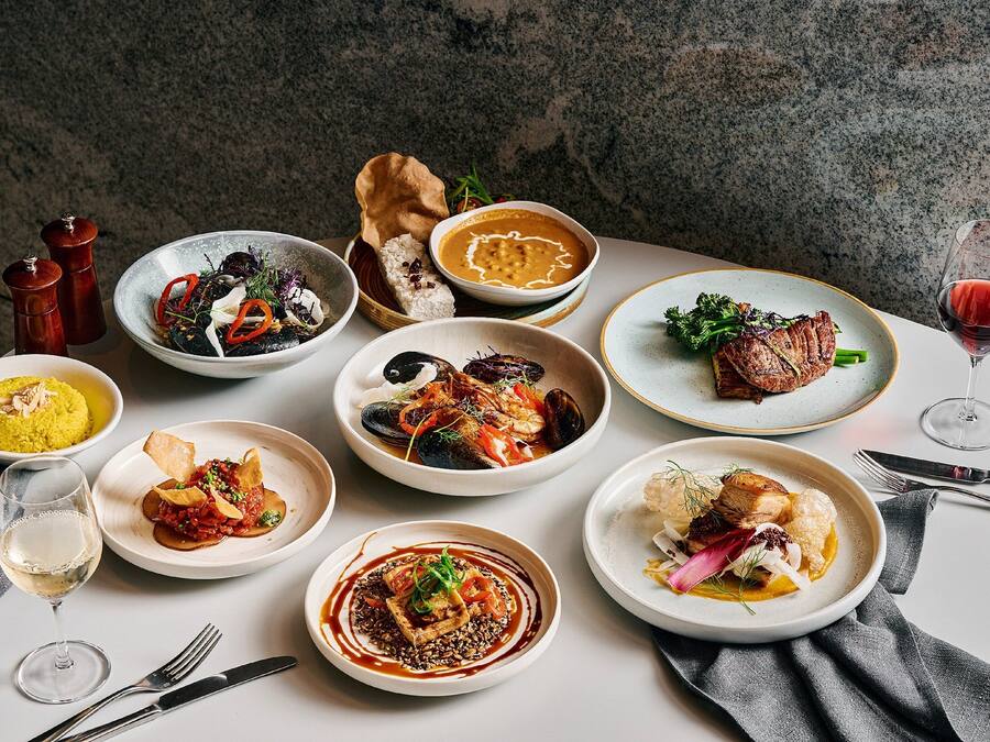 A variety of dishes on a table, including seafood, meat, and vegetables, with a wine glass and cutlery.