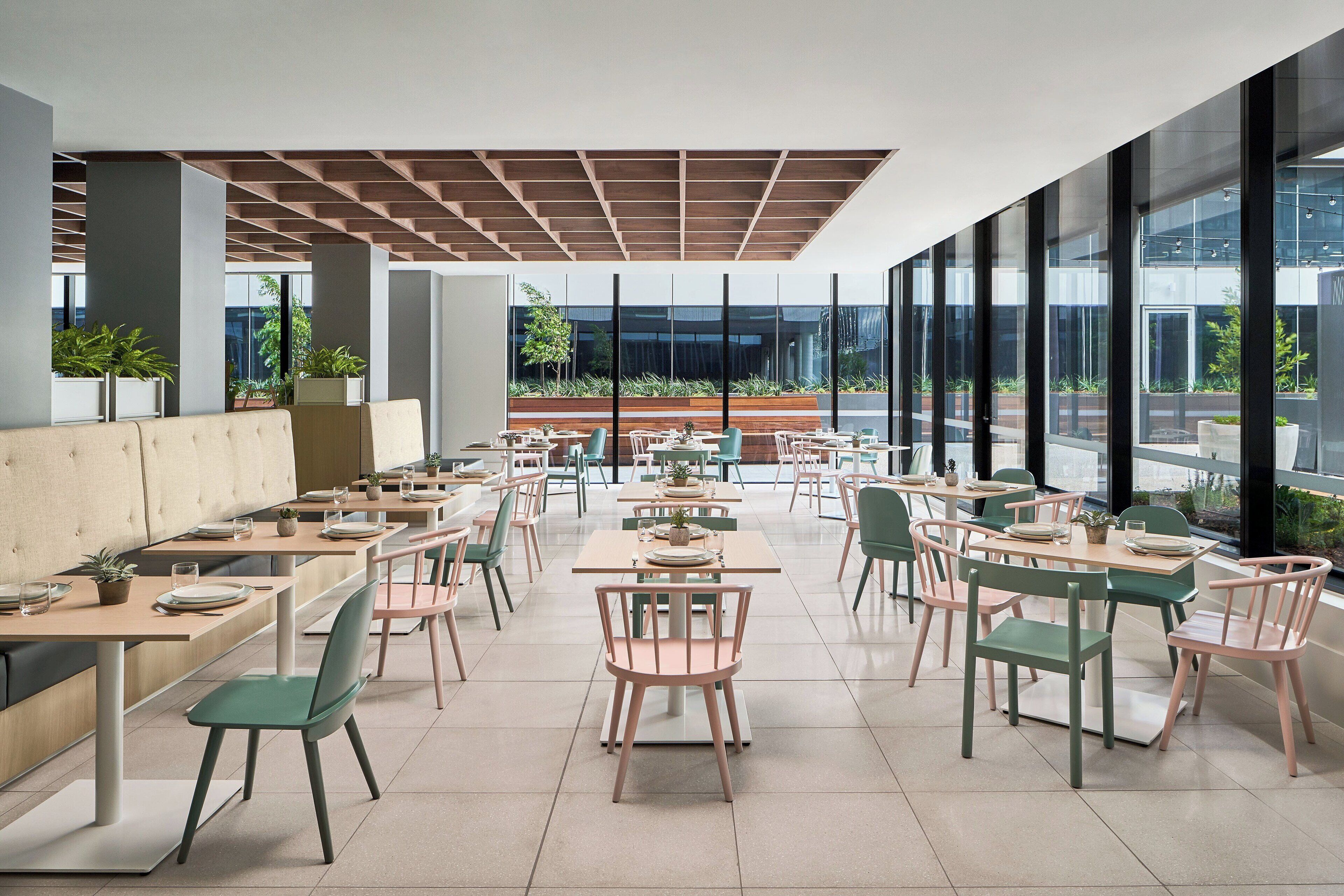 A modern restaurant with long communal tables and booths, large windows, and indoor plants.
