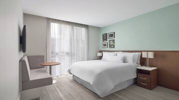 Une chambre d’hôtel avec un lit, un petit canapé, une table de chevet, une lampe et une fenêtre avec des rideaux légers.