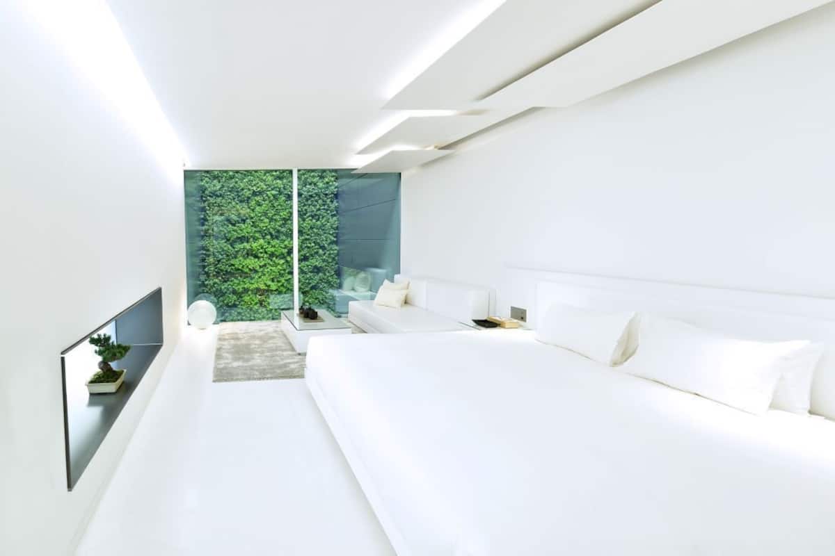 A modern bedroom with a large bed, a sofa, a TV, and a view of greenery through a glass wall.
