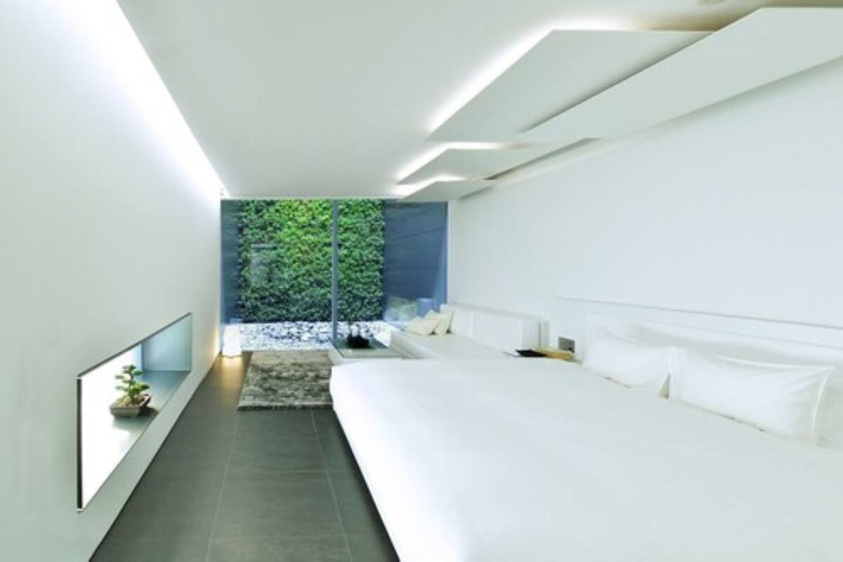A modern bedroom with a large bed, a small seating area, and a view of greenery through a glass wall.