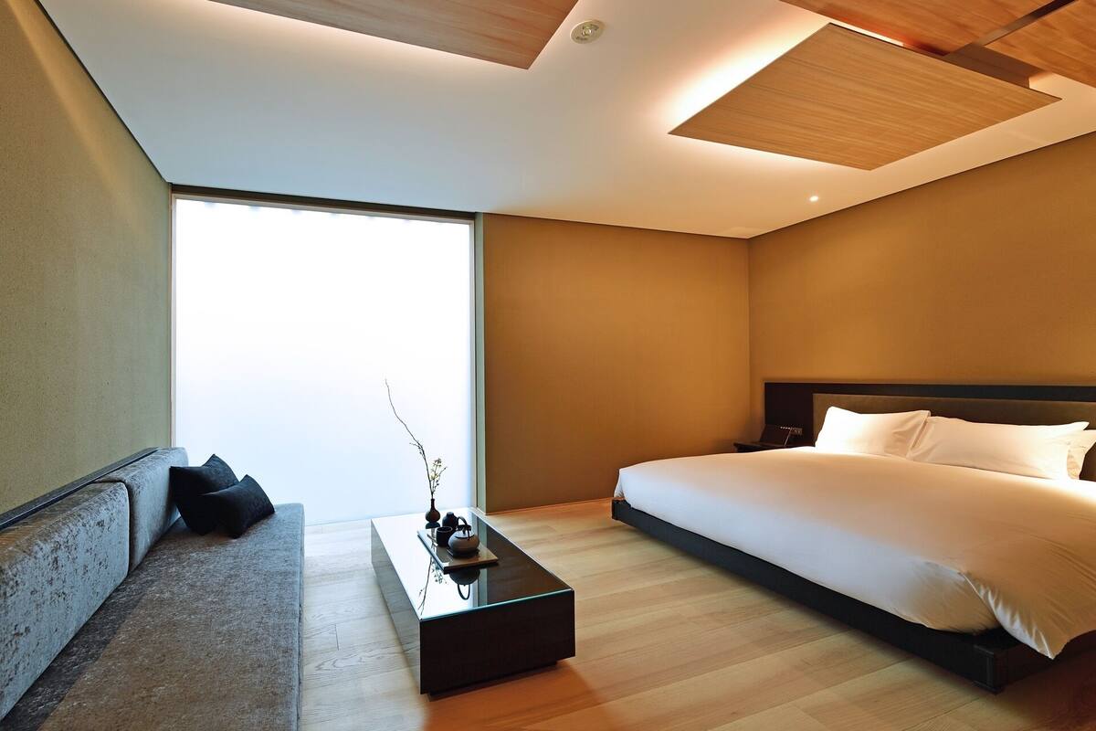 A modern hotel room with a large bed, a sofa, a coffee table, and a view of the sky.