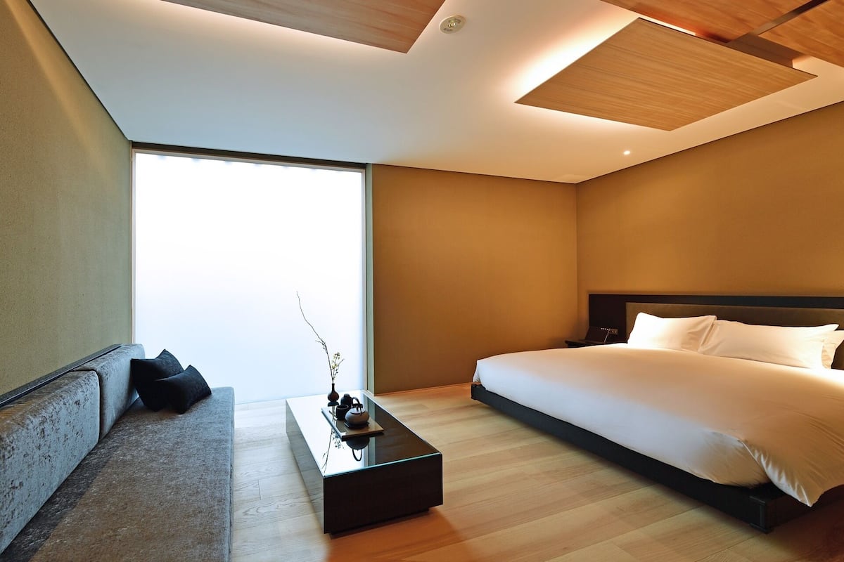 A modern hotel room with a large bed, a sofa, a coffee table, and a view of the sky.