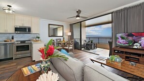 Ein modernes Wohnzimmer mit einer Couch, einem Couchtisch und einem Fernseher. Es gibt eine Küchenzeile mit einer Mikrowelle und einem Backofen. Das Zimmer verfügt über ein großes Fenster mit Blick auf den Ozean.