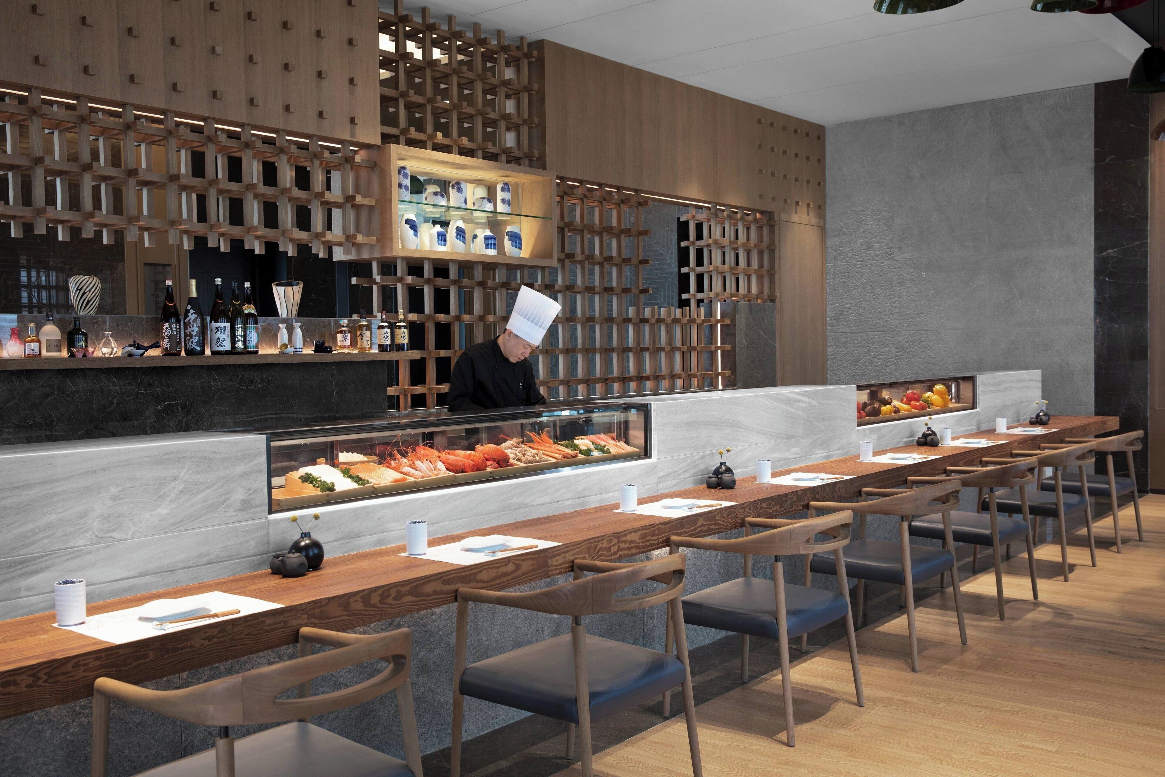 Un interior de restaurante moderno con un chef en la barra, sillas de madera y una vitrina con mariscos.