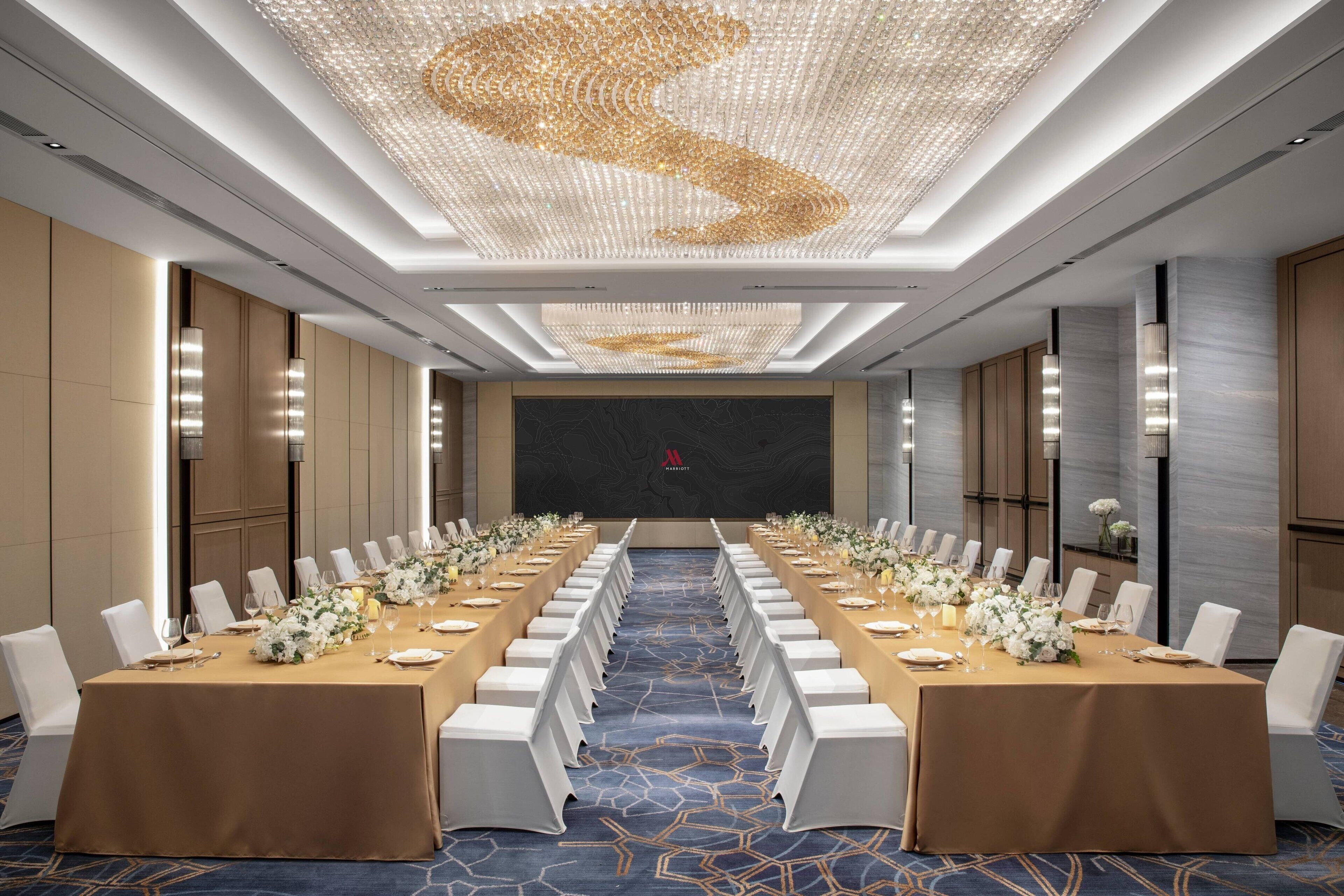 A conference room with a long table set for a meeting, a large screen at the front, and a decorative ceiling light.