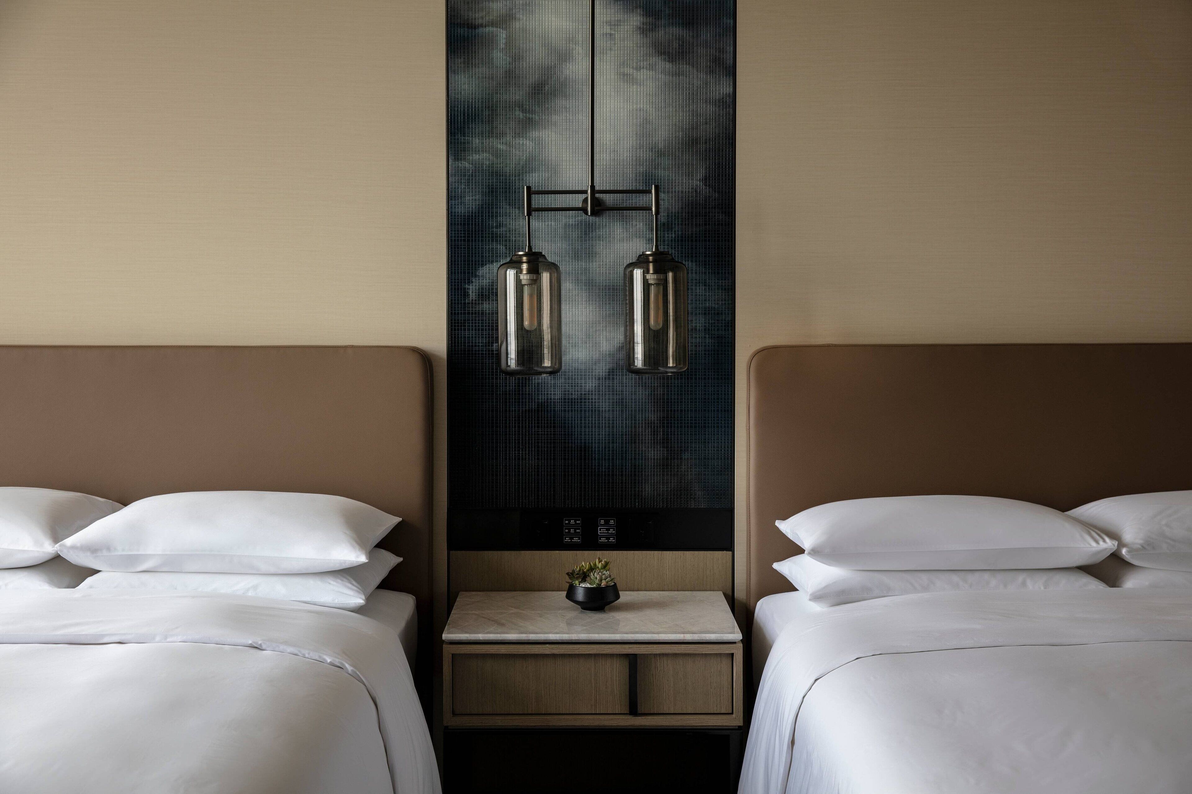 Quarto de hotel com duas camas, uma mesa de cabeceira com uma planta e um candeeiro fixo na parede.