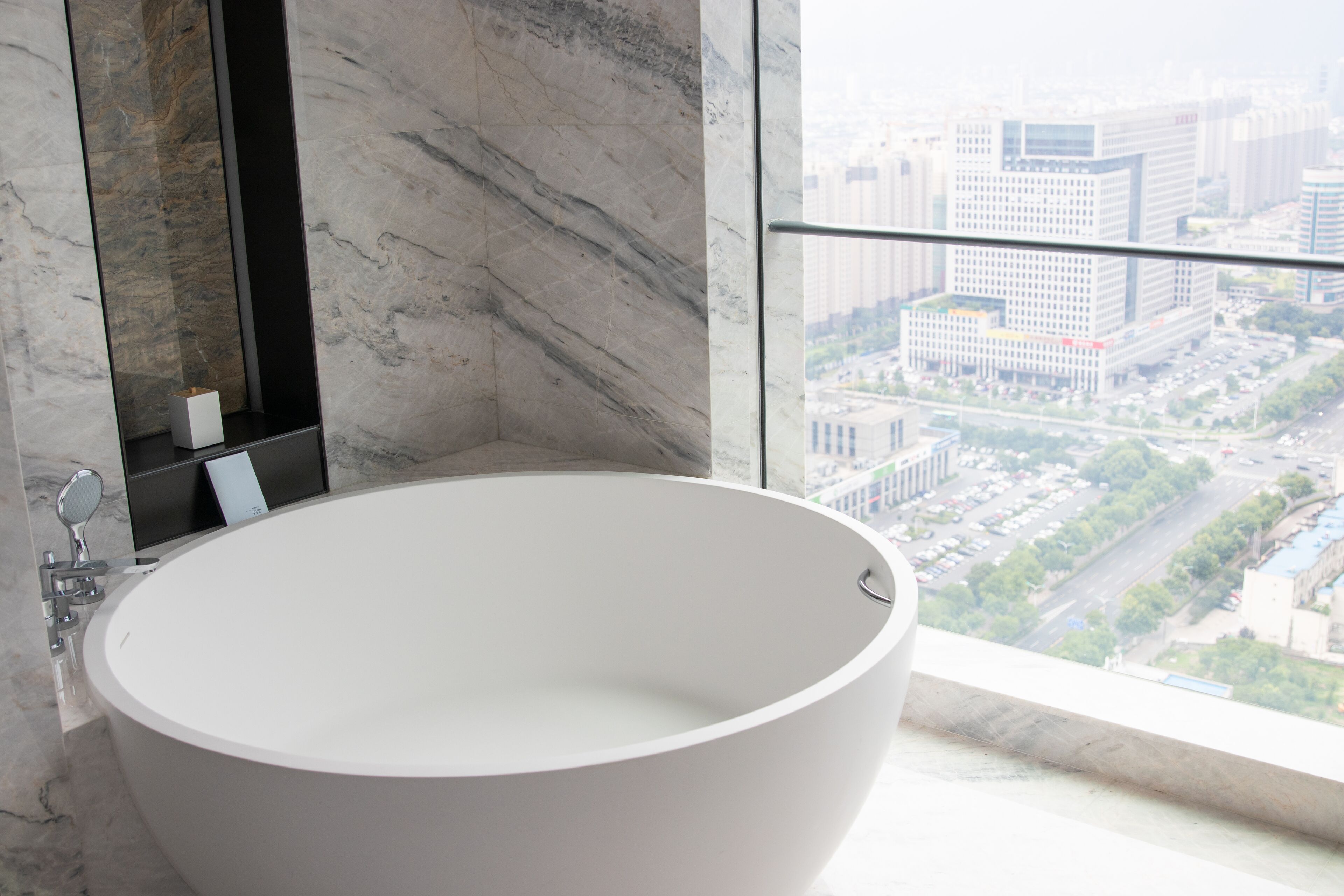 A modern bathroom with a freestanding bathtub, a marble wall, and a city view through a large window.