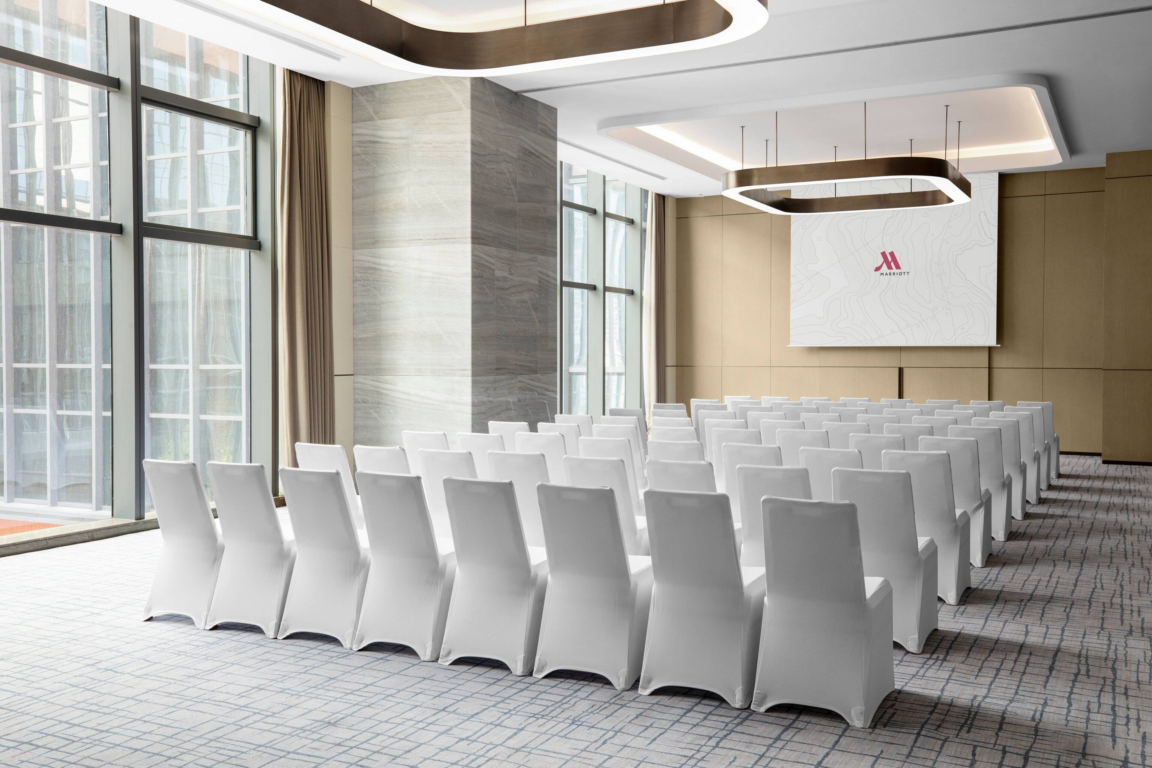 A conference room with rows of white chairs, large windows, and a modern ceiling design.