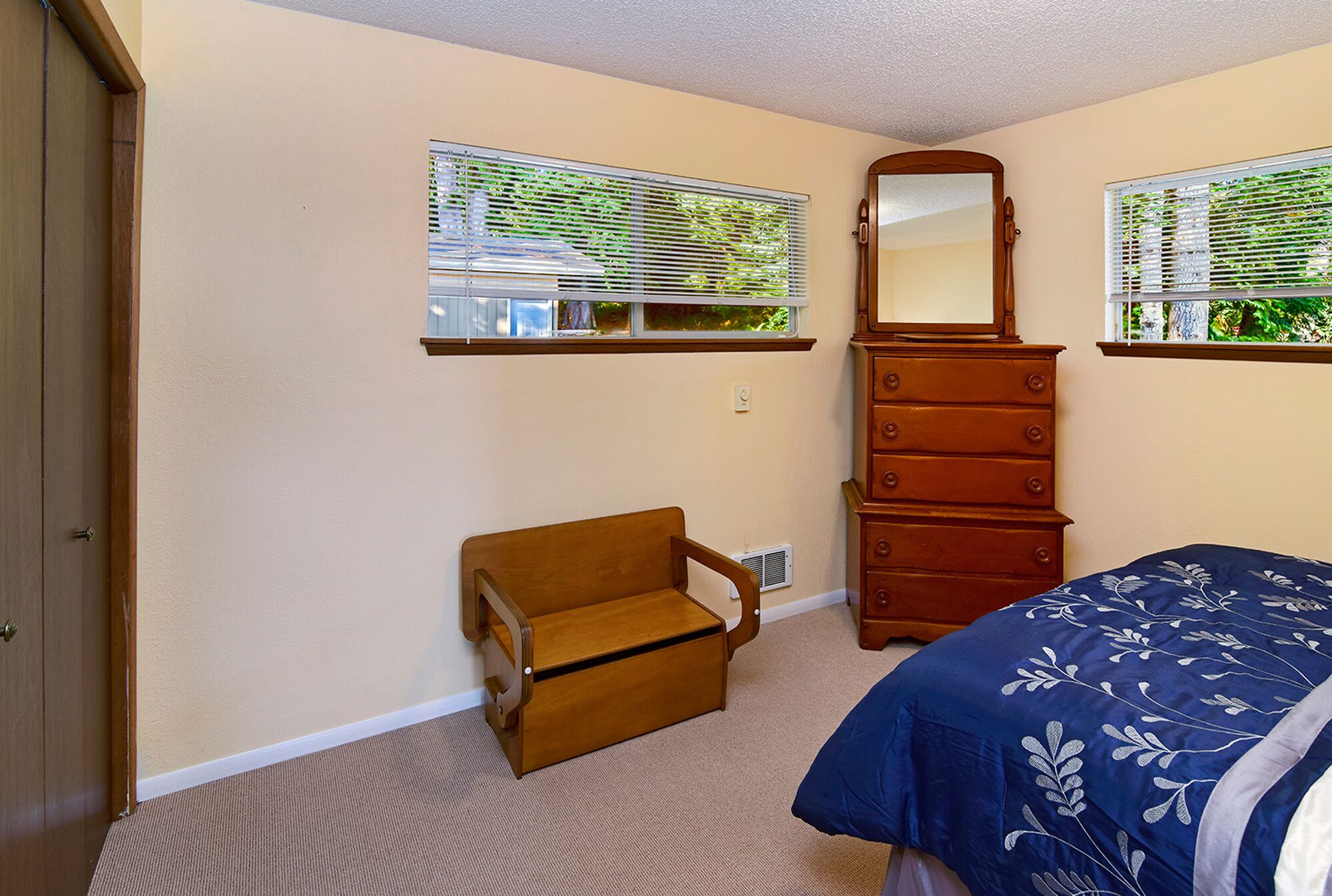Une chambre à coucher comprenant un lit, une commode, une chaise et deux fenêtres équipées de stores.