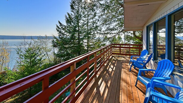 Eine Holzterrasse mit roter Geländer, Blick auf einen See, blaue Stühle und eine Ansicht von Bäumen sowie einer fernen Uferlinie.