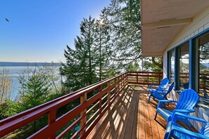 Una terraza de madera con barandilla roja que da a un lago, sillas azules y vistas a árboles y una orilla distante.
