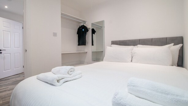 Habitación de hotel con una cama grande, ropa de cama blanca, cabecera gris y una puerta blanca.