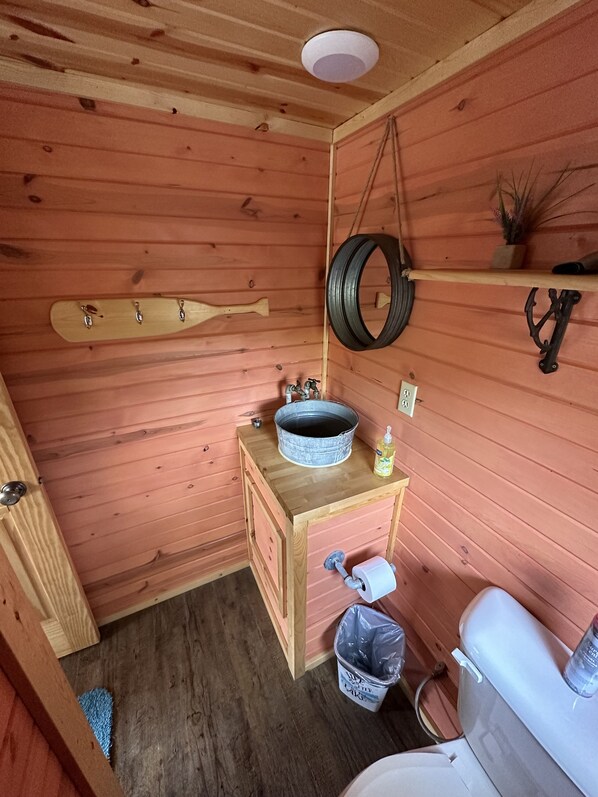 A compact wooden cabin bathroom with a wooden bench, a metal sink, a toilet, and a trash bin. - No Wake Zone (Jasonville)