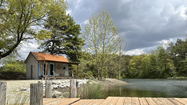 Une cabane rustique avec un toit en métal, une jetée s’avançant dans un lac, entourée d’arbres et d’un mur de pierres.