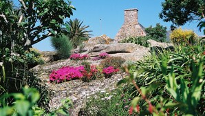 Un edificio de piedra con techo cónico, rodeado de abundante vegetación y plantas florecientes.