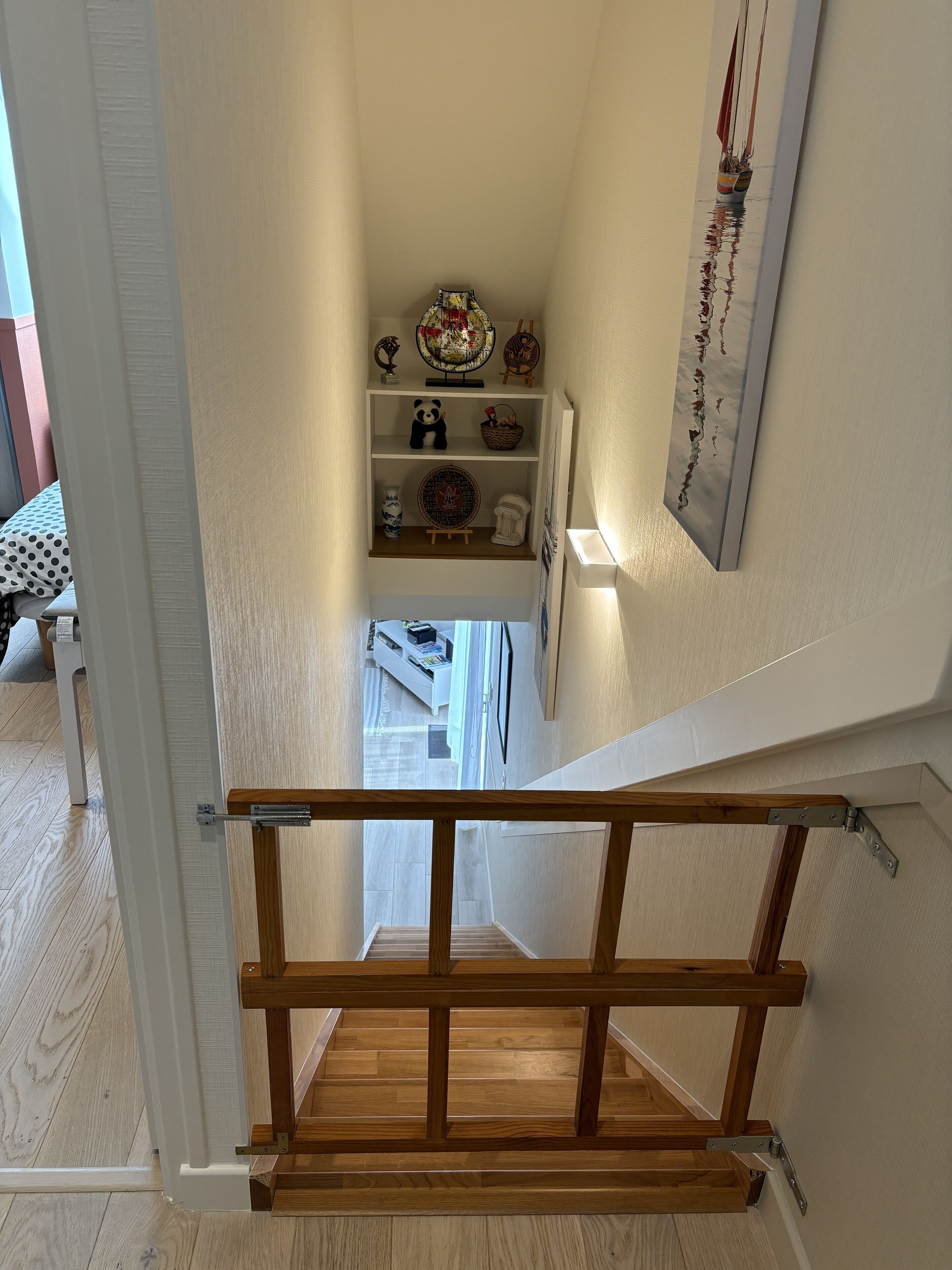 Un escalier en bois mène à un niveau inférieur, où l’on trouve un mur blanc et une étagère ornée d’objets décoratifs.