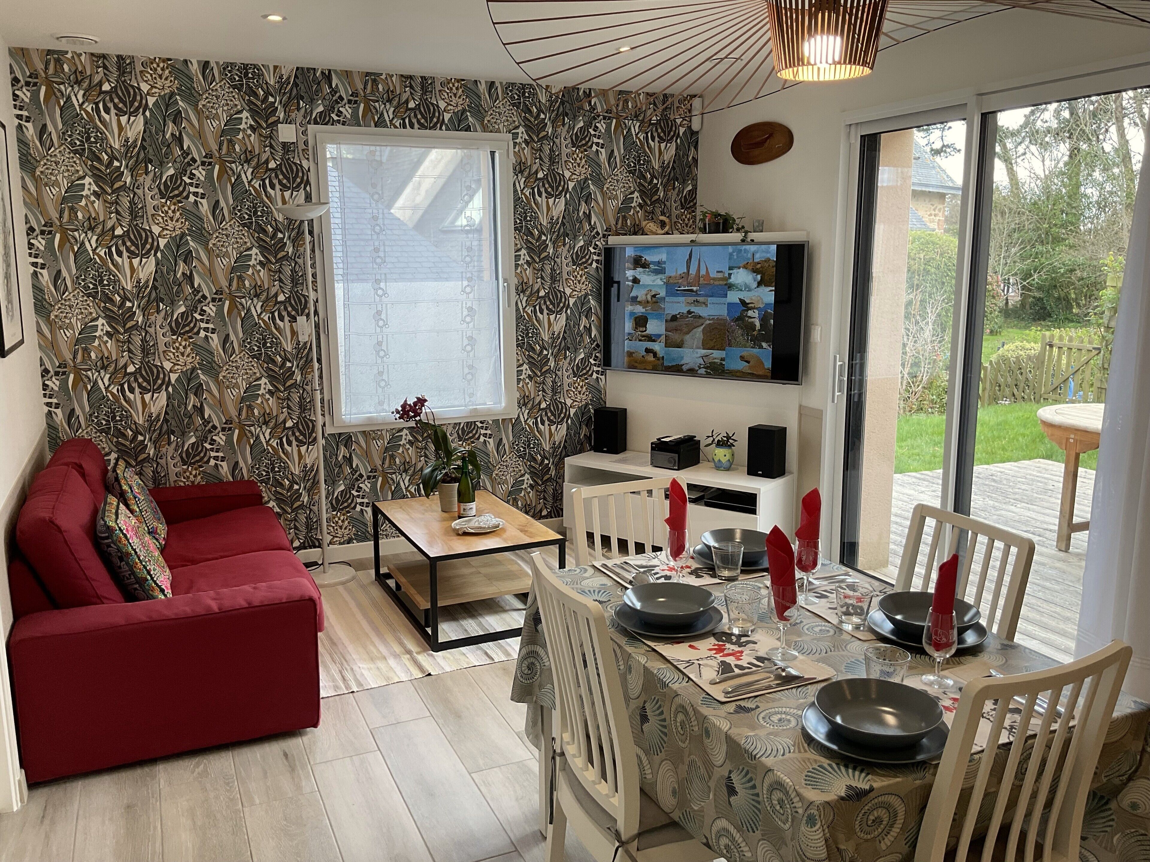 Un salon avec un canapé rouge, une table basse et un coin repas avec une table dressée pour quatre personnes. Un téléviseur est fixé au mur, et il y a une fenêtre donnant sur l’extérieur.