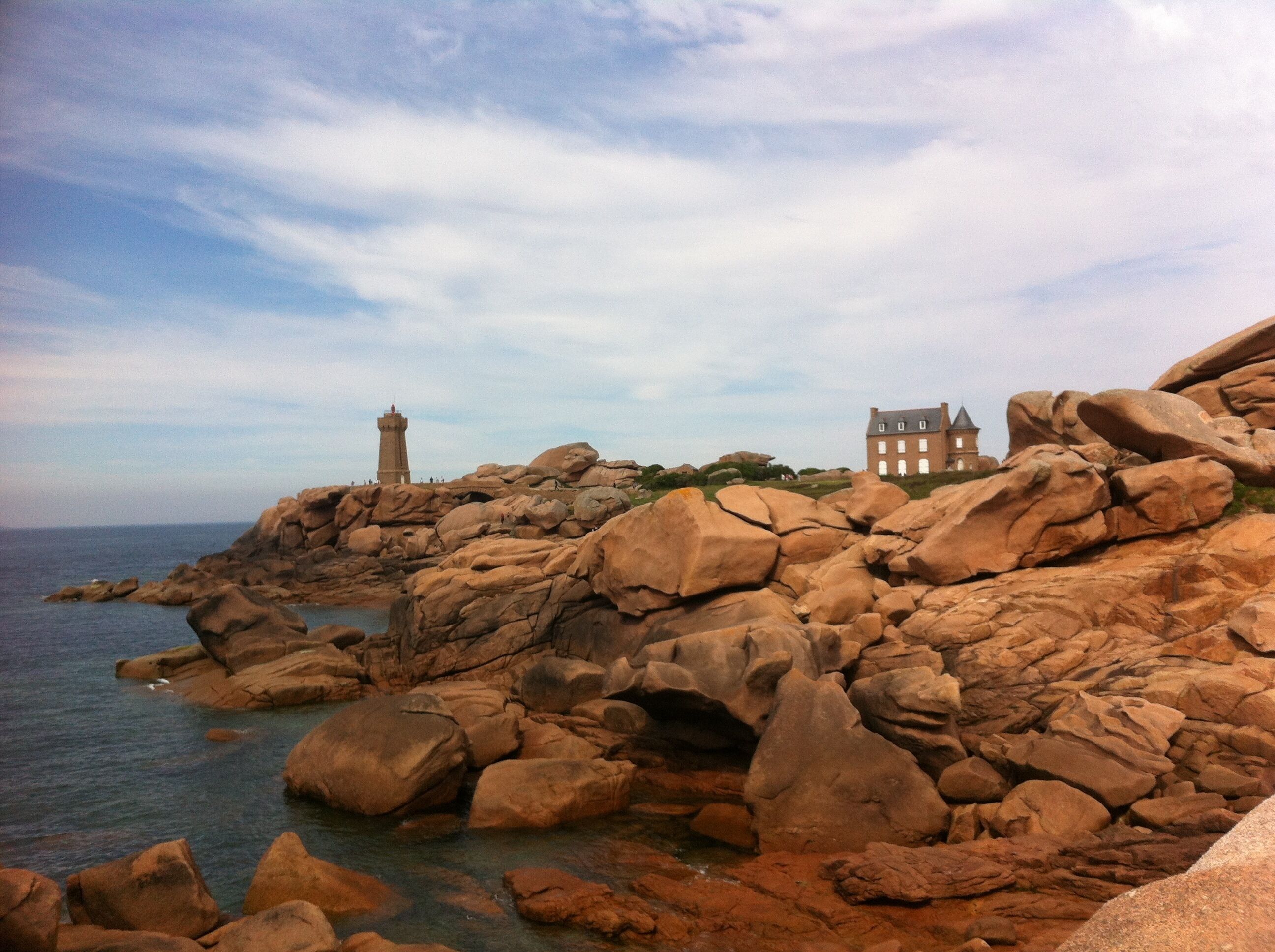 A coastal landscape with large boulders, a lighthouse, and a historic building.