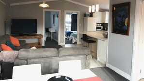 A living room with a grey sofa, a flat-screen TV, and a dining area with a red table. There is a kitchen with a microwave and a painting on the wall.