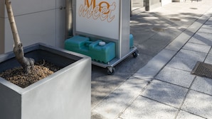 Um edifício moderno com área de serviço indicada, uma árvore em vaso e um carrinho com almofadas verdes.