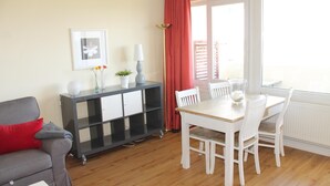 Uma sala de estar com um sofá cinza, uma mesa de jantar branca com cadeiras, um armário preto com gavetas, uma cortina vermelha e uma janela com persianas.