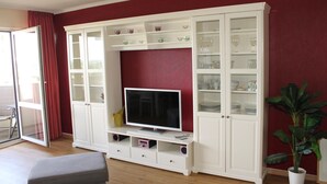 Sala de estar com televisão de tela plana em um rack branco, porta de vidro deslizante e planta em vaso.