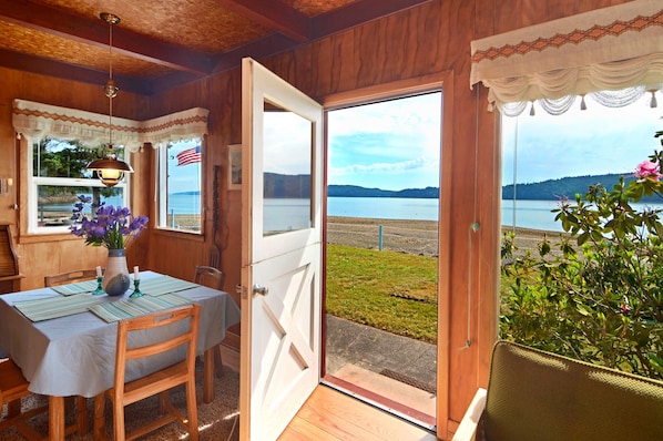 A wooden interior with a dining table set for two, a vase with flowers, and a view of a lake and mountains through an open door. - Charming little red beach house (Hoodsport)