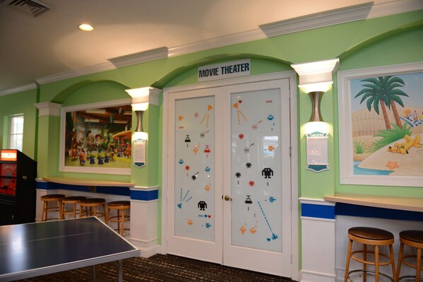 Une pièce avec une table de ping-pong, deux portes décorées de motifs rappelant les jeux d’arcade et une fresque murale représentant une scène de plage.