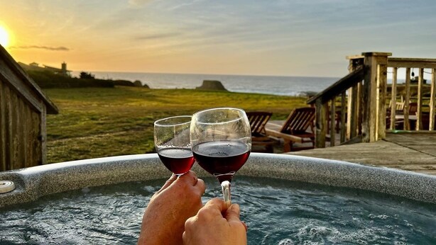 Een bubbelbad met twee personen die wijn glazen vasthouden, met uitzicht op een strand en oceaan.