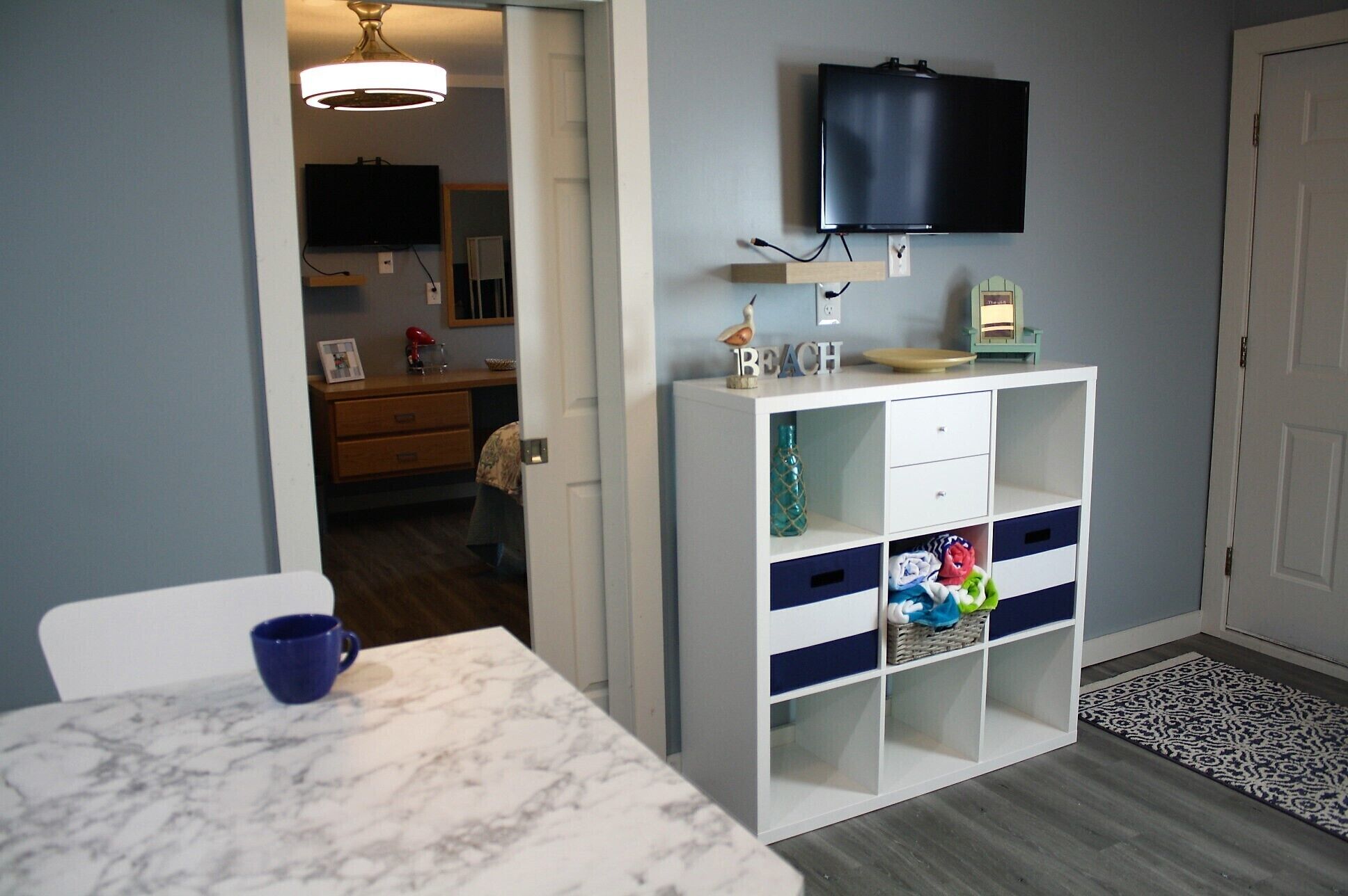 A compact kitchen with a marble countertop, a white cabinet with drawers, a television mounted above, and a small table with a blue cup.