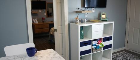 A compact kitchen with a marble countertop, a white cabinet with drawers, a television mounted above, and a small table with a blue cup.
