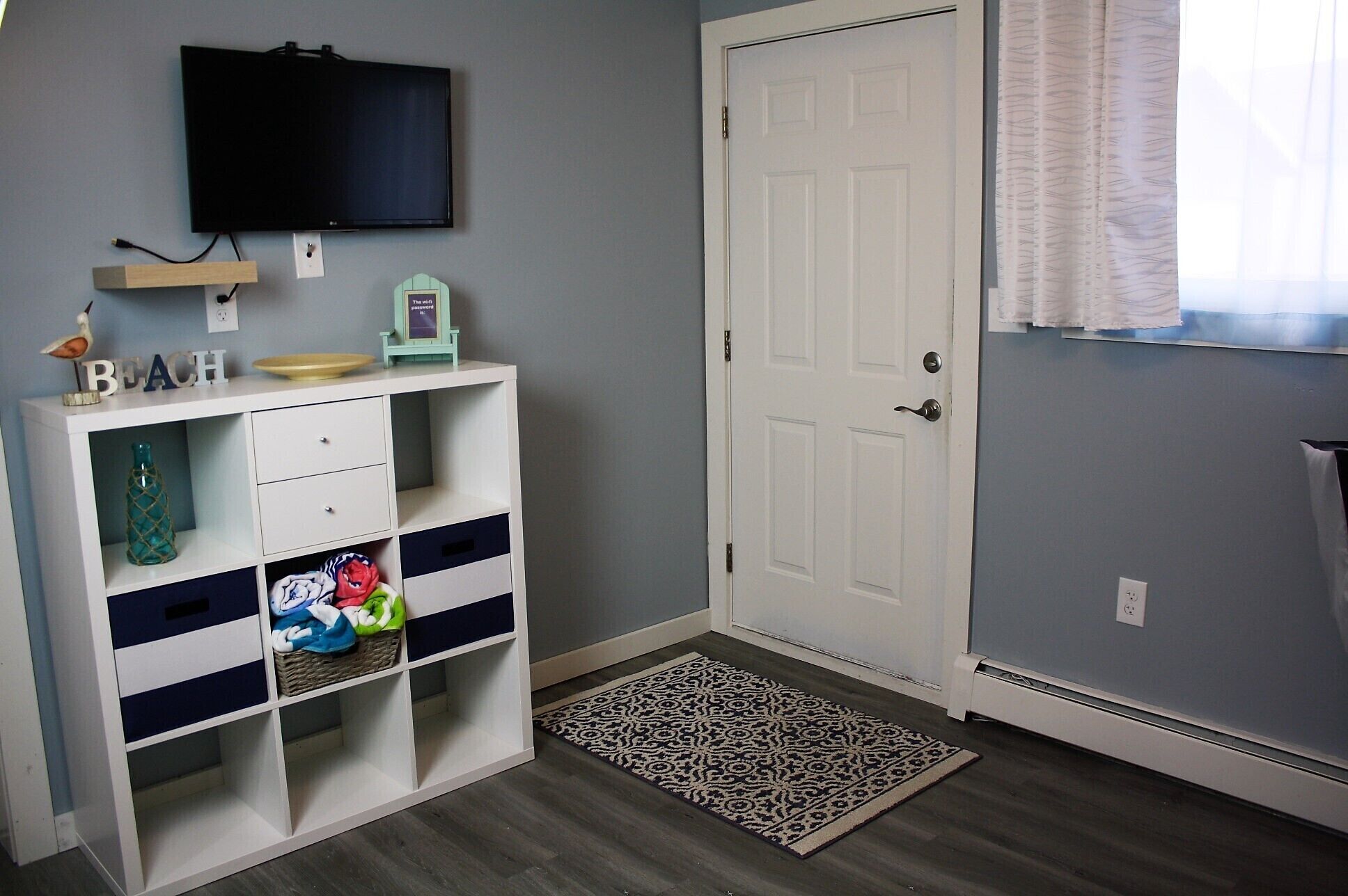 A room with a white door, a mounted TV, a shelf with decorative items, and a patterned rug.